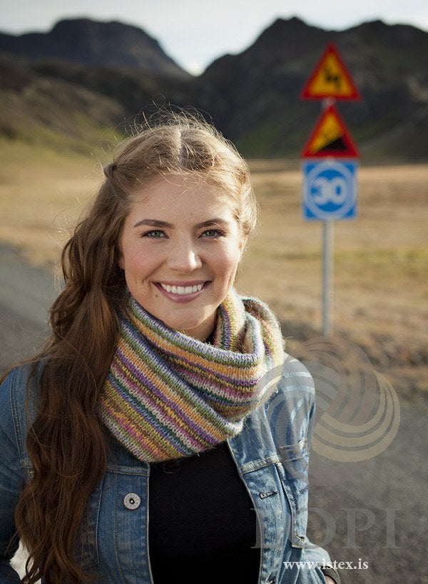 Woman wearing the Vindbelgur cowl wrapped twice round her neck — narrow, slightly slanting stripes in pink, yellow, purple, light blue-grey, green, teal, brown, straw and peach (shown in the original colours — this kit has mimosa for the sun yellow). The denim jacket and black top are not part of the pattern