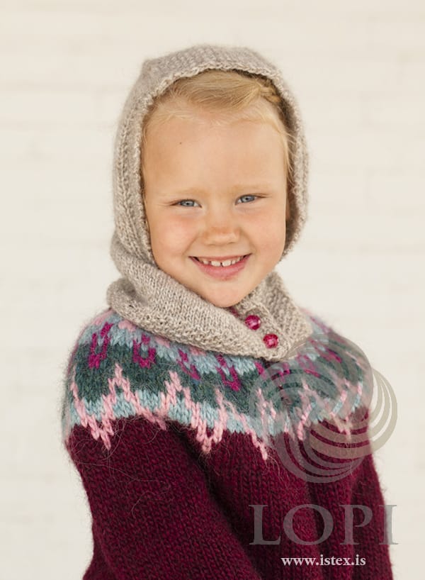 Girl wearing Hetta up as a hood in the pattern’s light beige heather, with dark pink buttons. Her burgundy sweater with a patterned yoke is a different pattern and not part of this one