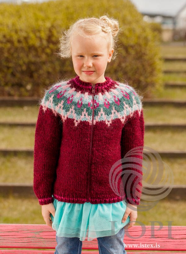 Girl standing in the red Kjós, zipped up to the collar, shown in the pattern’s own colours — oxblood red with a yoke in winter morning, arctic exposure, teal heather and dark magenta, and dark magenta stripes in the rib at the hem, cuffs and collar. The aqua tulle skirt, the jeans and the red bench are not part of the pattern