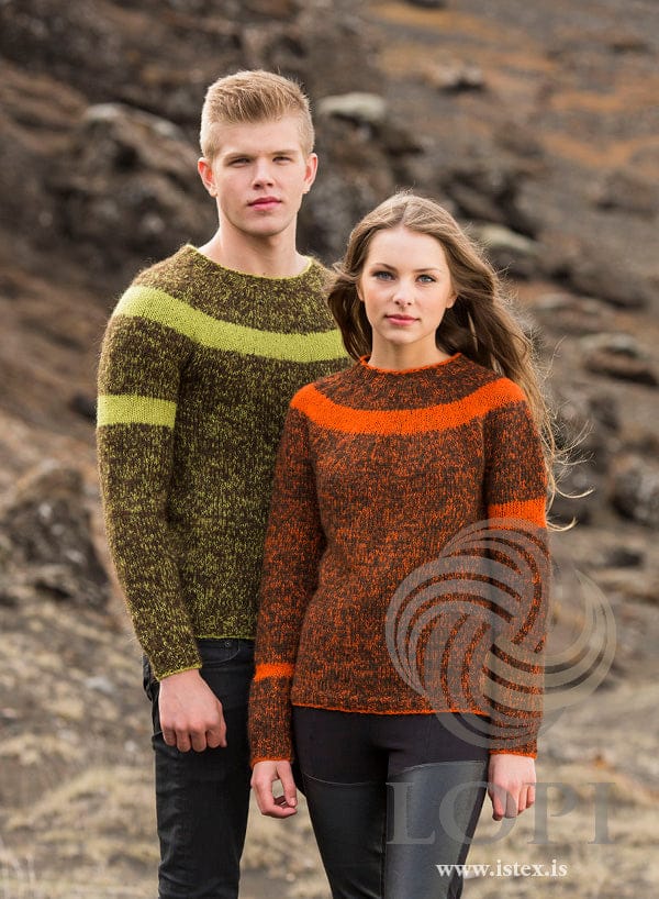 Young man and young woman on a hillside in the two Slydda versions, shown in the pattern’s own colours — his in lime Einband and chocolate Plötulopi, with a lime stripe round the yoke and on his right upper arm; hers in orange and chocolate, with an orange stripe round the yoke, on her left upper arm and near her right cuff. His black jeans and her black leggings with shiny panels are not part of the pattern