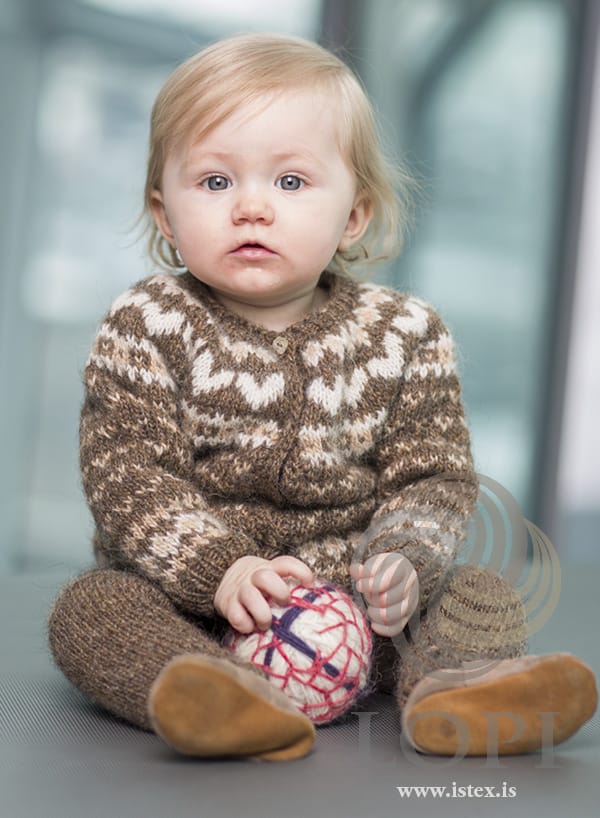 Baby sitting on the floor in Hjörtu, both pieces, shown in the pattern’s own colours — a buttoned murky (mid brown) cardigan with small straw V shapes, a straw and white band above the hem and cuffs and a row of big white hearts round the yoke, and plain murky trousers. The ball and the tan suede shoes are not part of the pattern