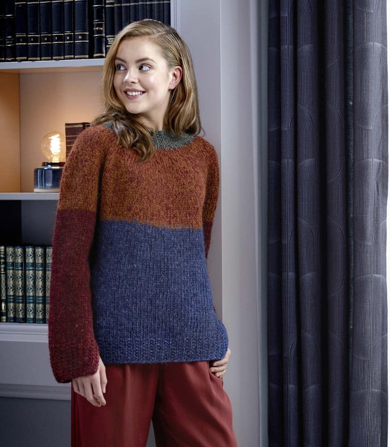 Woman by a bookcase in the Fletir sweater — a blue lower body with a deep ribbed hem, deep red sleeves with long ribbed cuffs, a rust-amber yoke over the top of the body and sleeves, and a dark grey-green neckband (shown in the original colours — this kit has forest green for the discontinued forest in the neckband, so the neckband will look greener). The rust-red trousers are not part of the pattern
