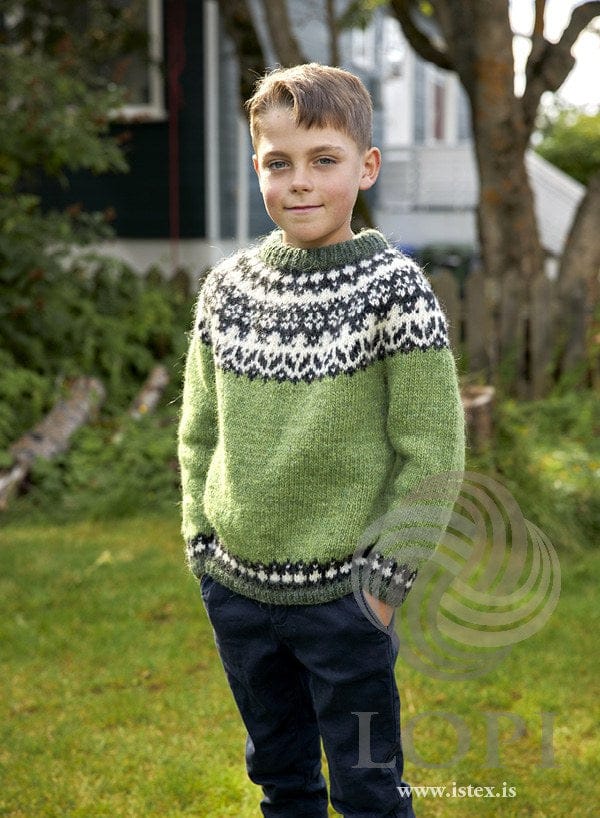Boy in a garden wearing the Ómur sweater — spring green with a galaxy and white yoke, and a lyme grass rib and neckband