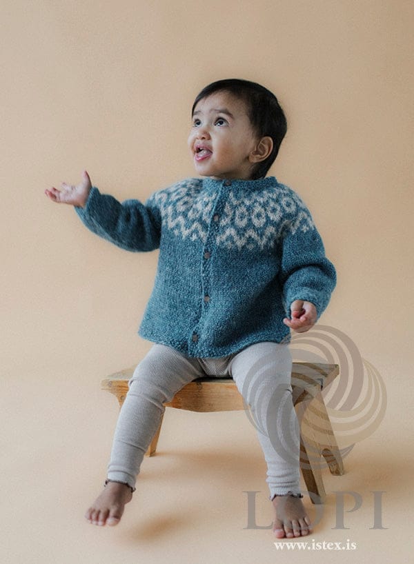 Toddler sitting on a small wooden stool in Tryggð — a gulf stream (teal blue) baby cardigan, buttoned up, with ivory beige zigzags and ovals round the yoke and seed-stitch edges at the hem, cuffs, neck and front bands, shown in the pattern’s own colours. The ribbed leggings and the stool are not part of the pattern