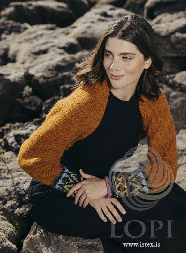 Woman sitting on mossy lava in the apricot Armur shrug, with multicoloured cuffs on galaxy