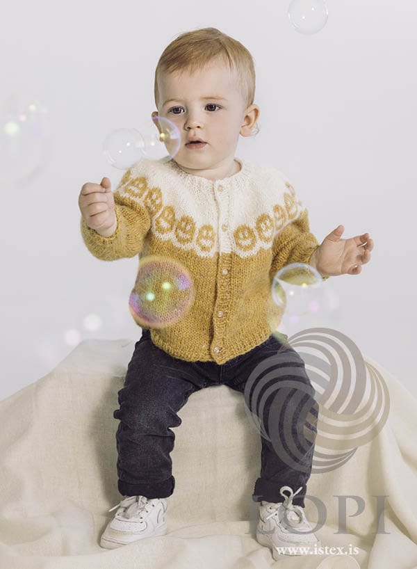 Toddler reaching for soap bubbles in Bros, buttoned — golden yellow body and sleeves, a white yoke with a row of smiling faces, and front bands that are yellow on the body and white on the yoke, in the pattern’s own colours. The dark jeans, the white trainers, the cream cloth and the soap bubbles are not part of the pattern