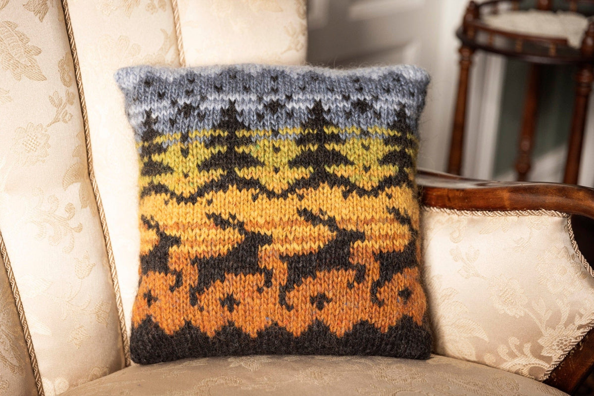 Hjörð cushion knitted in Álafosslopi — black reindeer and fir trees against bands of amber, golden, green and blue