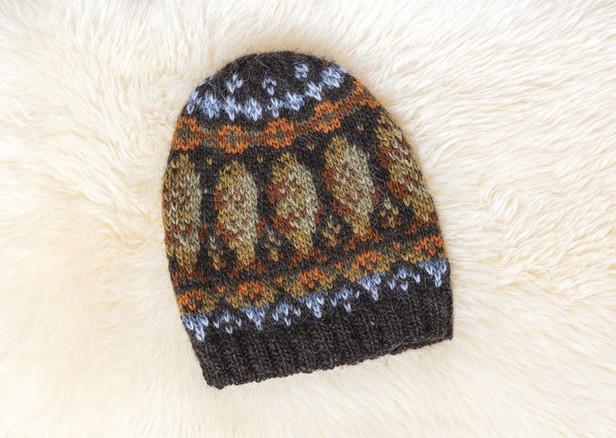 Lofoten Hat laid flat on a sheepskin: black sheep heather rib brim, light blue and apricot bands, and a band of large frostbite green and golden ovals with a rust checker inside